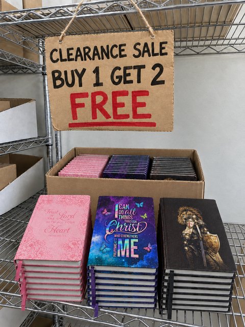 A cardboard sign on our warehouse shelf: Clearance sale, buy 1 get 2 free, above boxes of Blessac prayer journals.