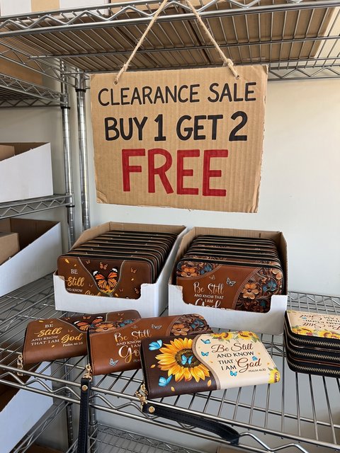 A cardboard sign on our warehouse shelf: Clearance sale, buy 1 get 2 free, above boxes of Blessac wallets.
