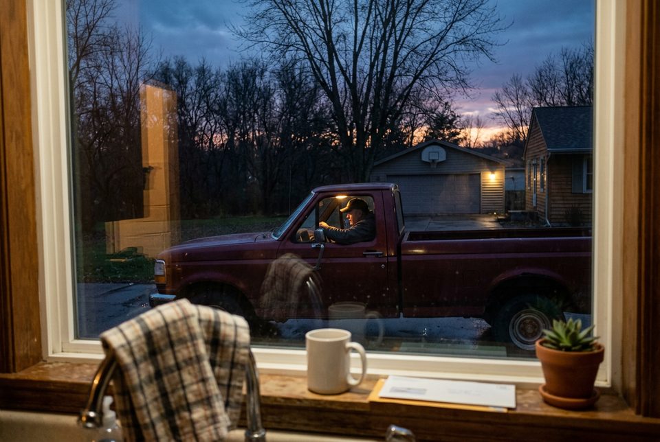 A pickup truck in a driveway at dusk, seen through a kitchen window, a man sitting still behind the wheel